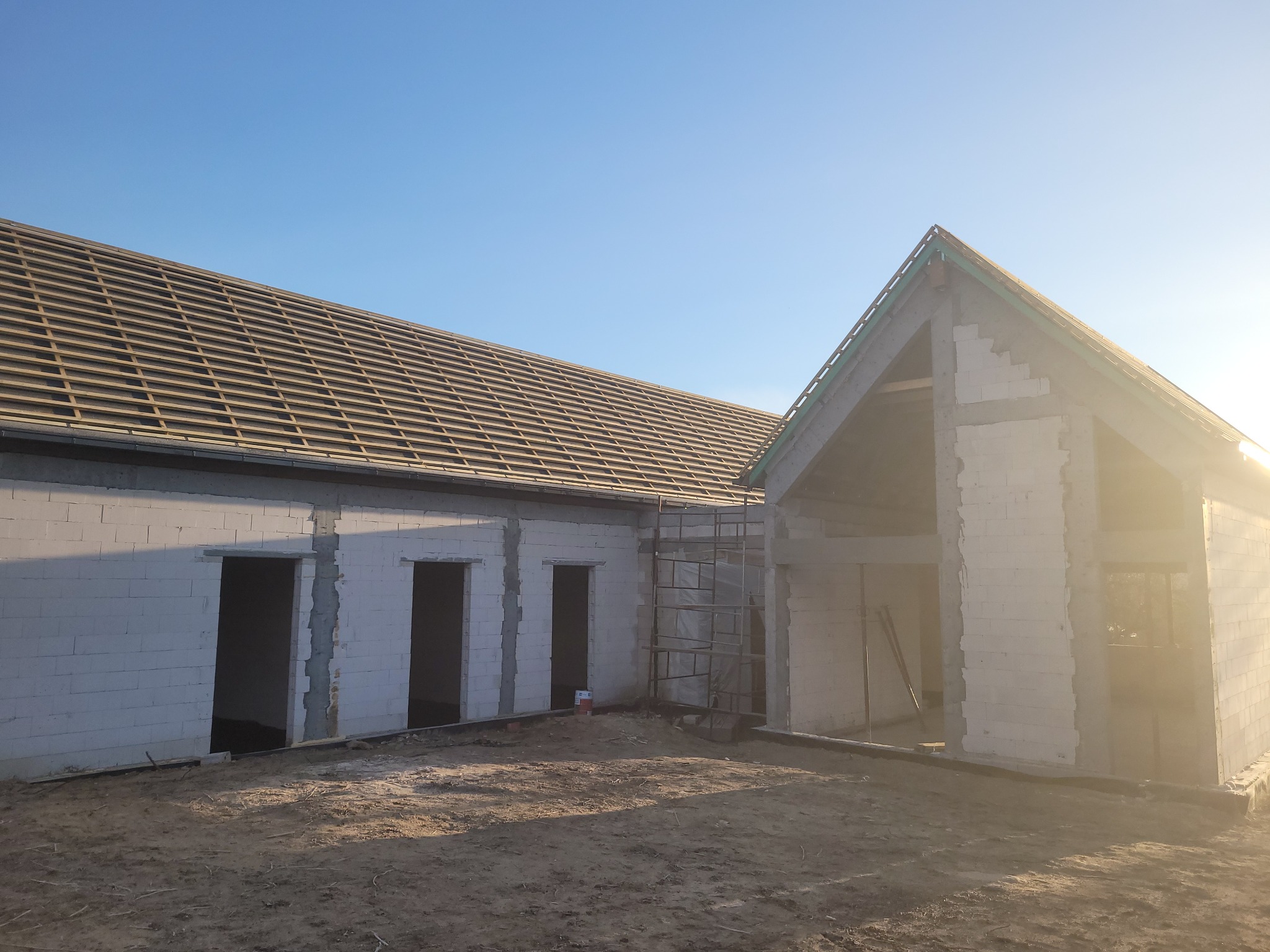 Surowy stan domu z białych bloczków, widoczne otwory okienne i drzwiowe, więźba dachowa w trakcie montażu, na pierwszym planie ziemia.