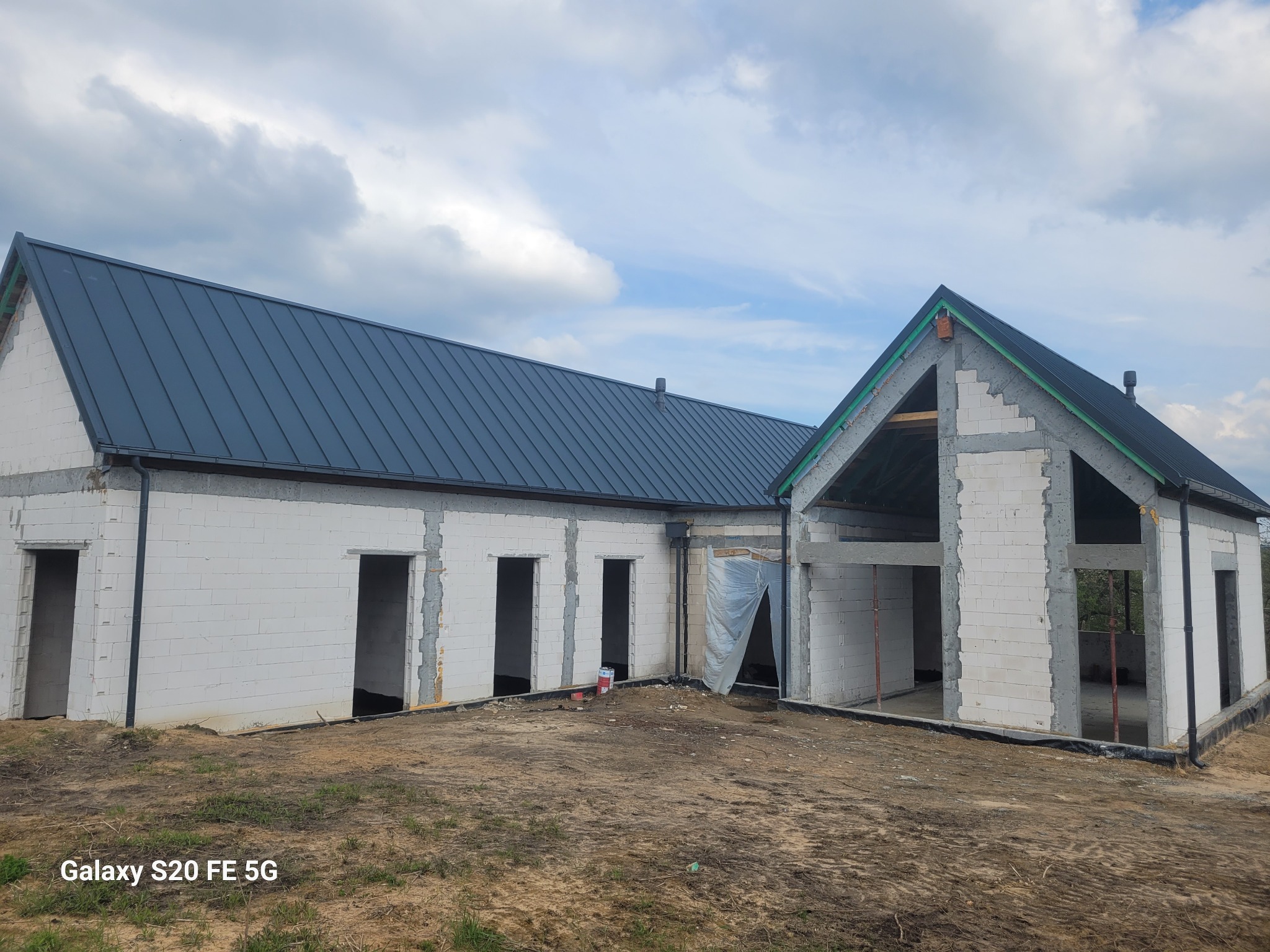 Surowy, biały dom z dachem z blachy trapezowej w trakcie budowy. Widoczne otwory okienne i drzwiowe, teren wokół w stanie naturalnym, pochmurne niebo w tle.