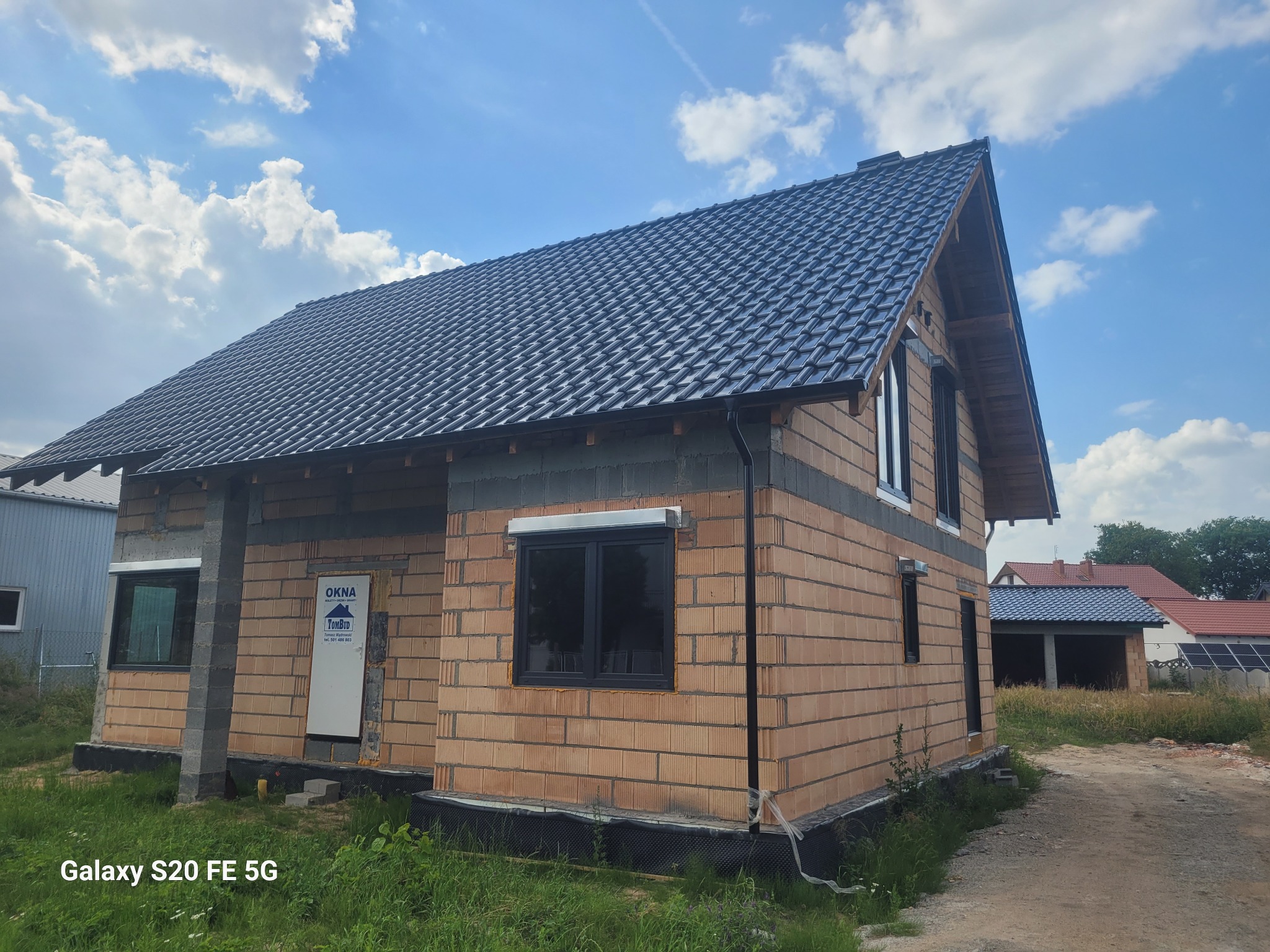 Dom w trakcie budowy z czarnym dachem i oknami. Widoczne bloczki ceramiczne, brama z logo firmy. W tle błękitne niebo z chmurami.
