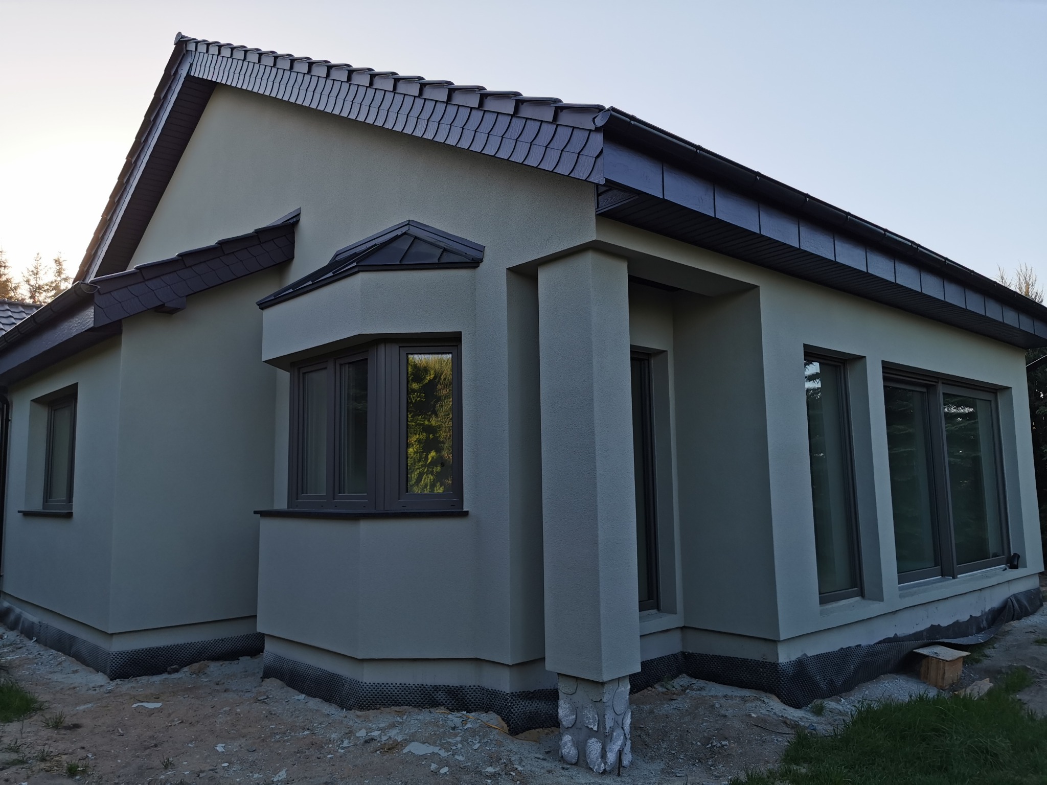 Nowoczesny dom z szarą elewacją i ciemną dachówką, widok z zewnątrz, okna z ciemnymi ramami, teren wokół domu w trakcie prac wykończeniowych.