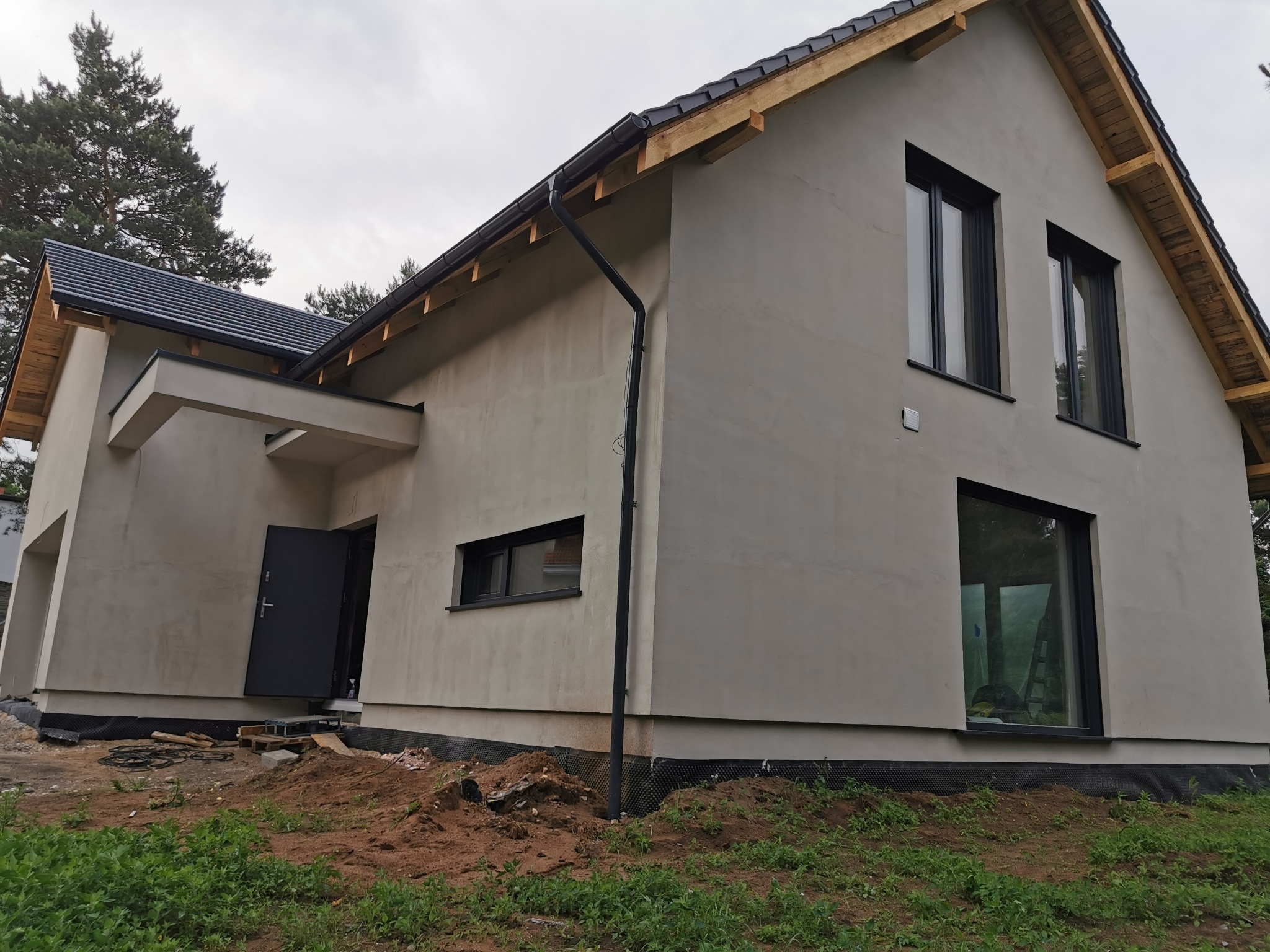 Nowoczesny dom jednorodzinny w trakcie wykończenia elewacji, z widocznym orynnowaniem i stolarką okienną w kolorze antracytowym, na tle pochmurnego nieba.