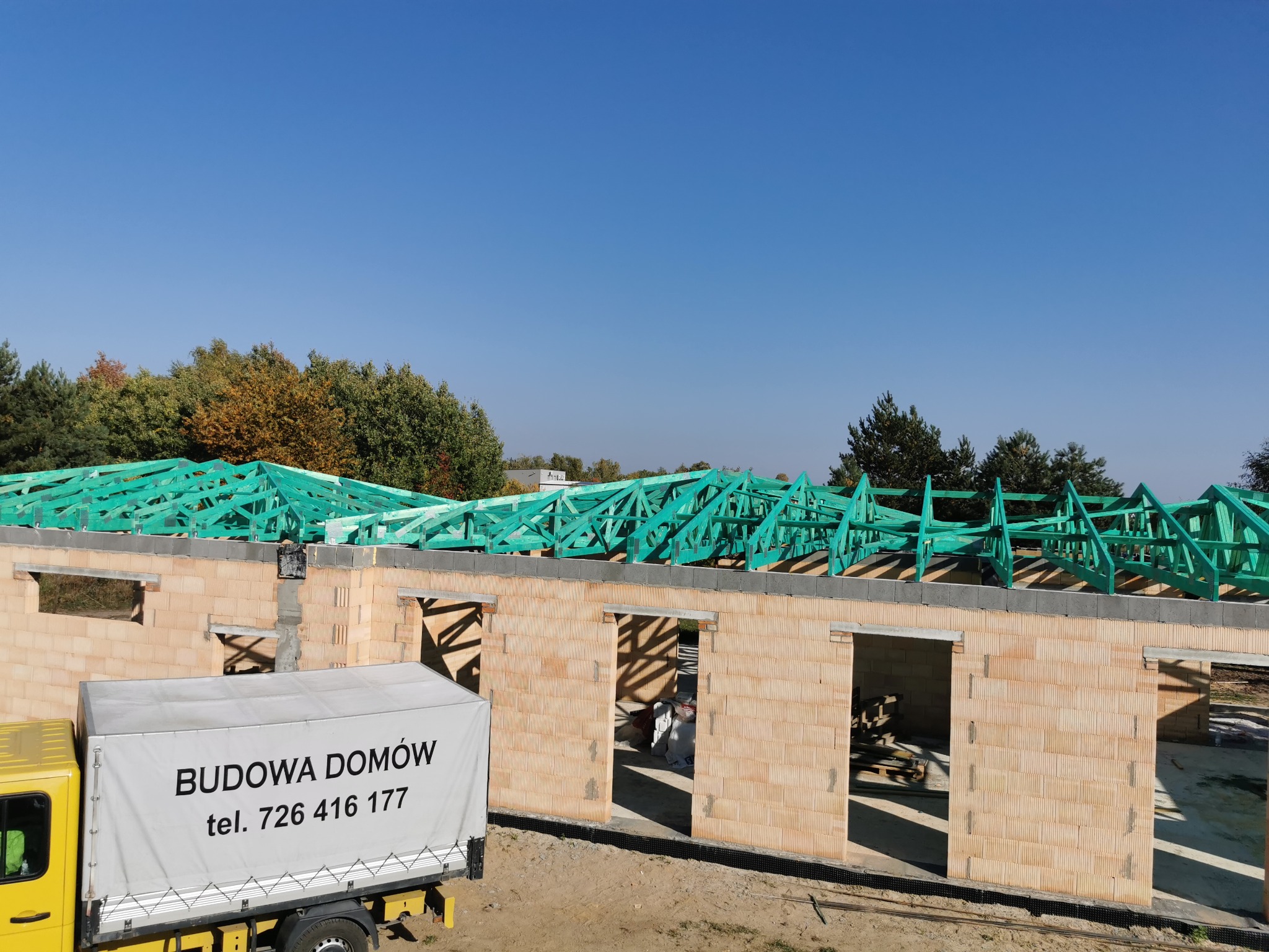 Wznoszony dom z pustaków ceramicznych, z widoczną konstrukcją dachu z impregnowanego drewna. Na pierwszym planie samochód dostawczy z napisem 'Budowa Domów'.