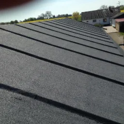 Szary dach pokryty papą termozgrzewalną z widocznymi łączeniami, w tle zabudowania gospodarcze i drzewa.