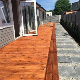 Nowoczesny taras z desek o ciepłym, brązowym odcieniu, przylegający do budynku z szarą elewacją. Obok ścieżka z szarych płyt betonowych oddzielonych białym żwirem, prowadząca wzdłuż betonowego...