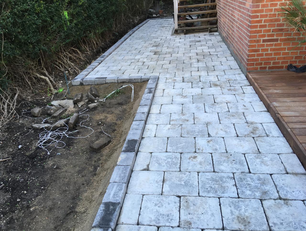 Ścieżka z jasnoszarej kostki brukowej, obrzeża z ciemniejszej kostki, widoczne przygotowane podłoże z kablami i kamieniami, w tle budynek z cegły i drewniany taras.