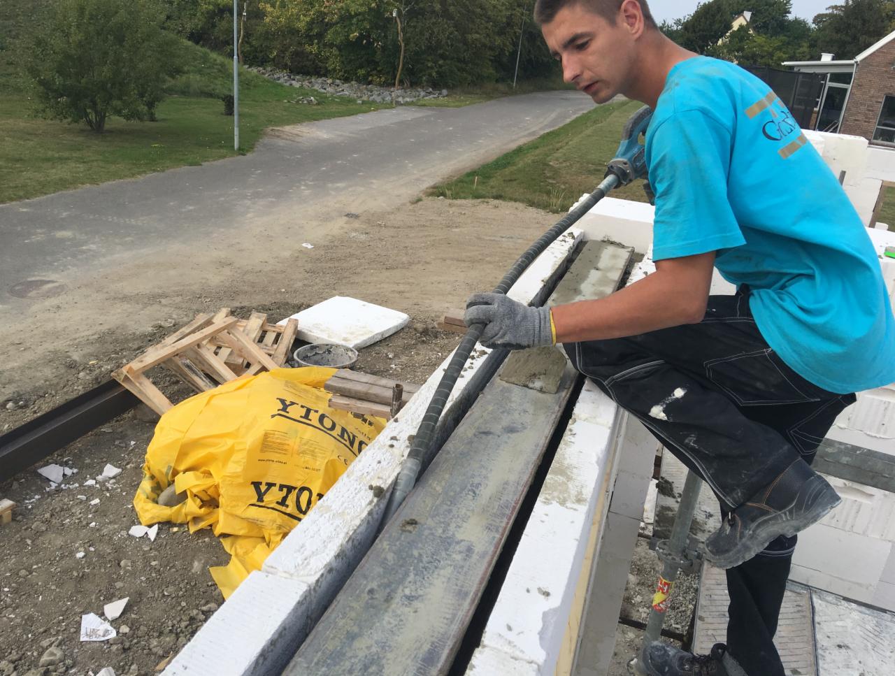 Pracownik w niebieskiej koszulce aplikuje zaprawę do spoin między bloczkami z betonu komórkowego YTONG, widoczne elementy konstrukcyjne budynku w trakcie budowy.