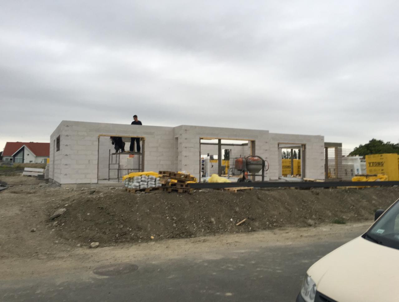 Budowa domu z bloczków Ytong: widoczne ściany w stanie surowym, palety z materiałem, betoniarka i pracownicy na rusztowaniu.