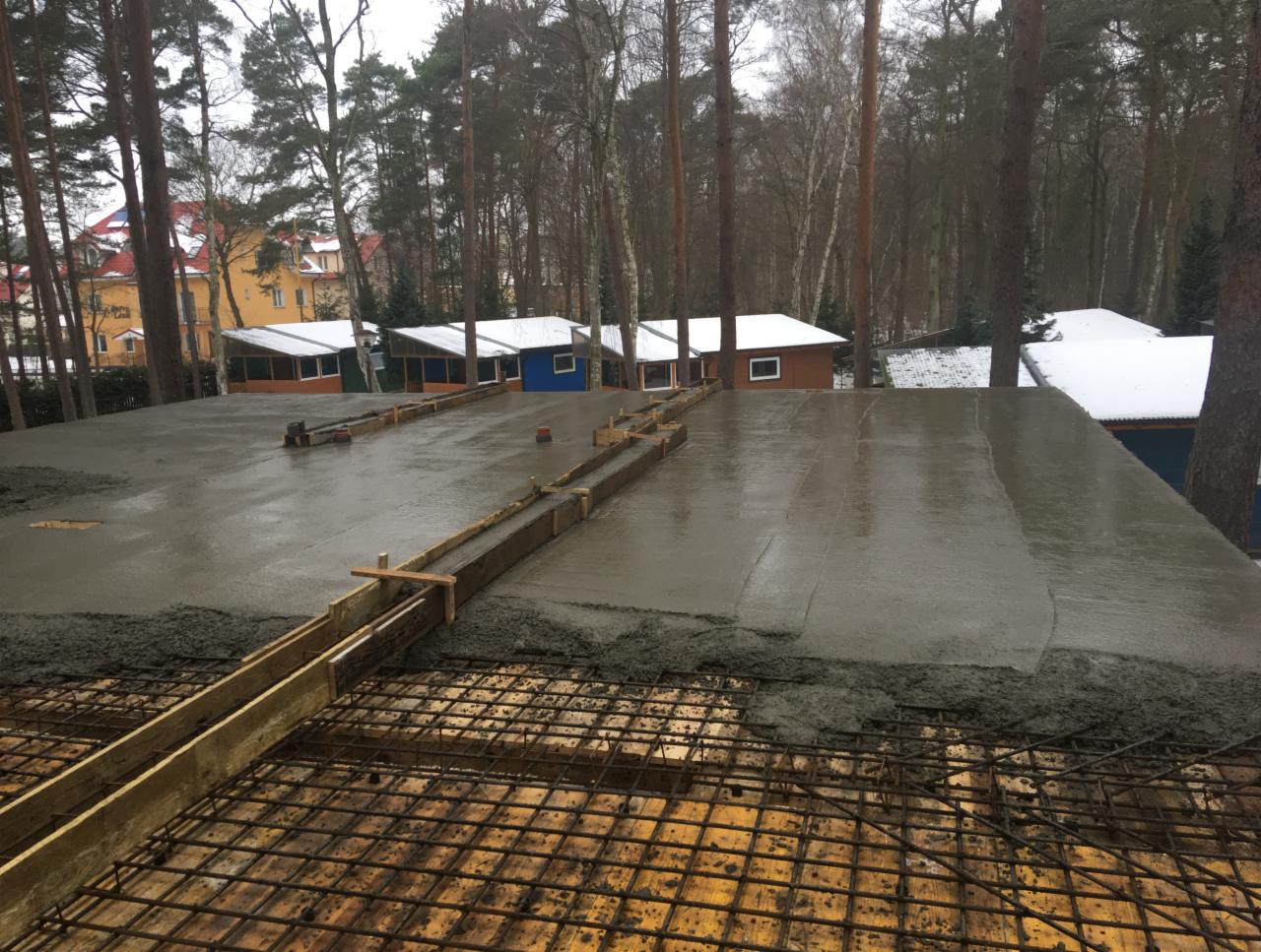 Wylewanie betonu na zbrojenie, szalunki drewniane, w tle budynki i drzewa.