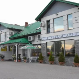 Budynek 'Dom Weselny Arkadia' z zielonym dachem, klimatyzacją i kamienną elewacją. Widoczne okna odbijające niebo i rośliny w donicach przed budynkiem.