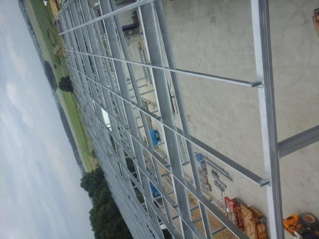 Stalowa konstrukcja hali w trakcie budowy, widok z góry, zieleń w tle. Elementy konstrukcyjne w kolorze srebrnym, maszyny budowlane widoczne w oddali.