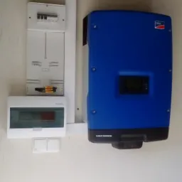 Niebieski falownik SMA Sunny Tripower zamontowany na białej ścianie obok skrzynki z bezpiecznikami i przewodami elektrycznymi.