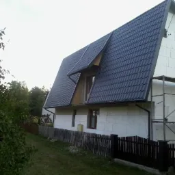 Dom w trakcie budowy z nowym, ciemnym dachem z dachówki, ocieplony styropianem, z rusztowaniem przy ścianie bocznej.