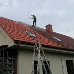 Pracownik w zielonej koszulce czyści czerwoną dachówkę myjką ciśnieniową, stojąc na dachu dwupiętrowego domu z beżową elewacją, widoczne okna dachowe i drabina oparta o budynek.