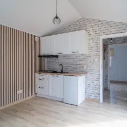 Wnętrze domu modułowego: aneks kuchenny z białymi szafkami, czarną armaturą, ściana z cegły i drewniane panele ścienne, widok na kolejne pomieszczenie przez otwarte drzwi.