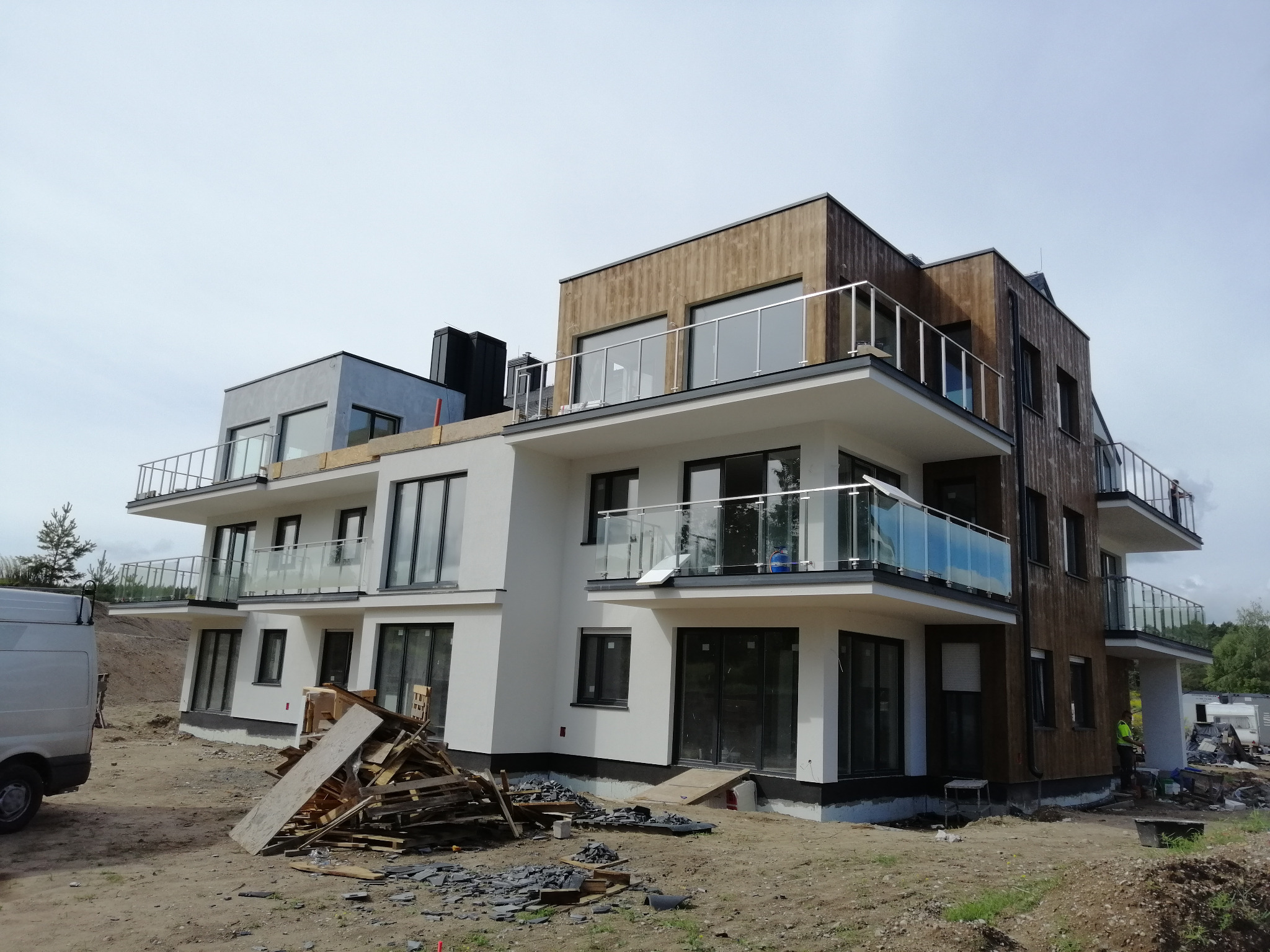 Dwupiętrowy budynek w trakcie budowy z drewnianą fasadą i balkonami z szklanymi balustradami, widoczne materiały budowlane i samochód dostawczy.