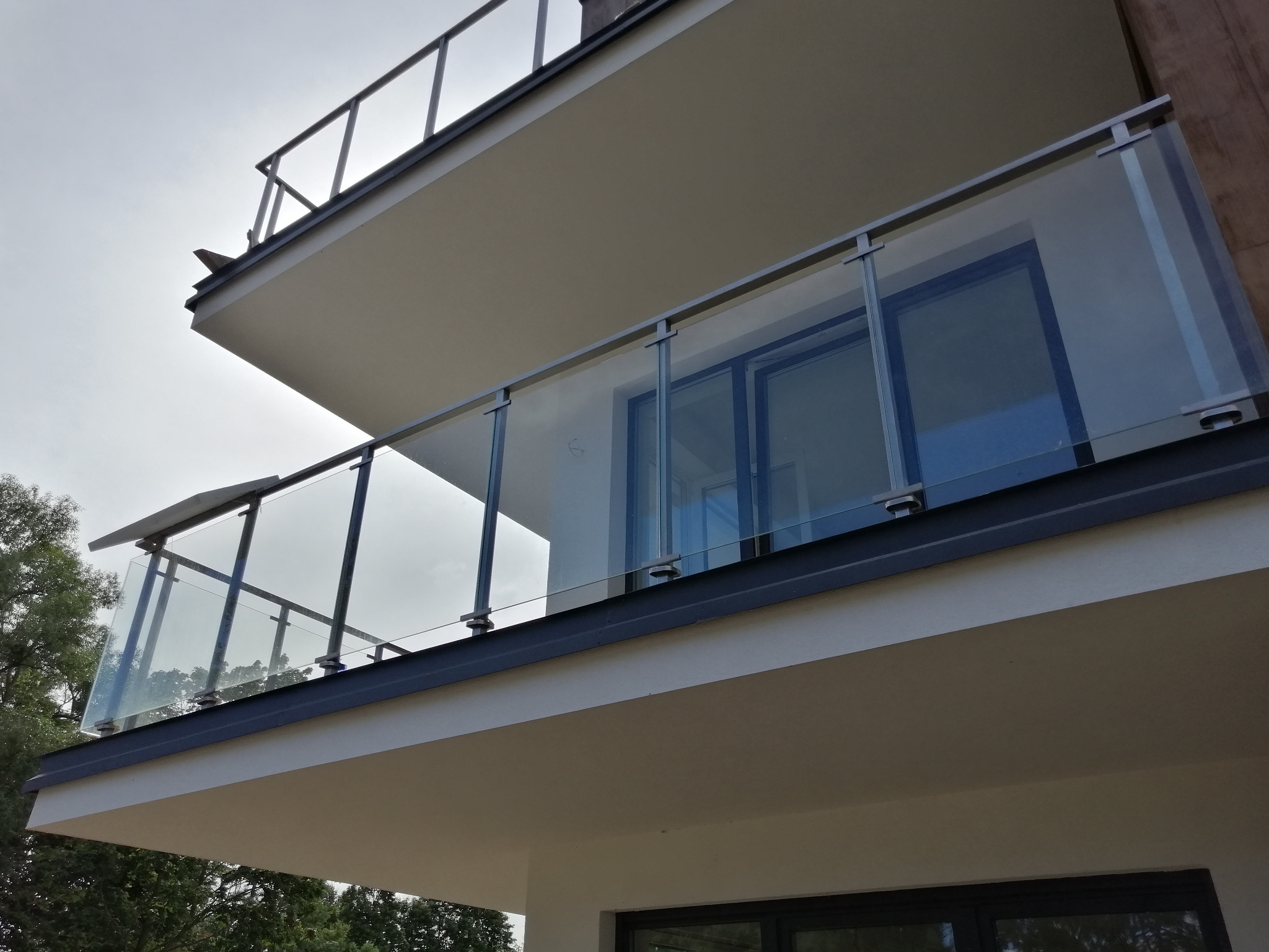 Nowoczesny balkon z szklaną balustradą i metalowymi elementami, widok z dołu na dwupiętrowy budynek z drzewami w tle.