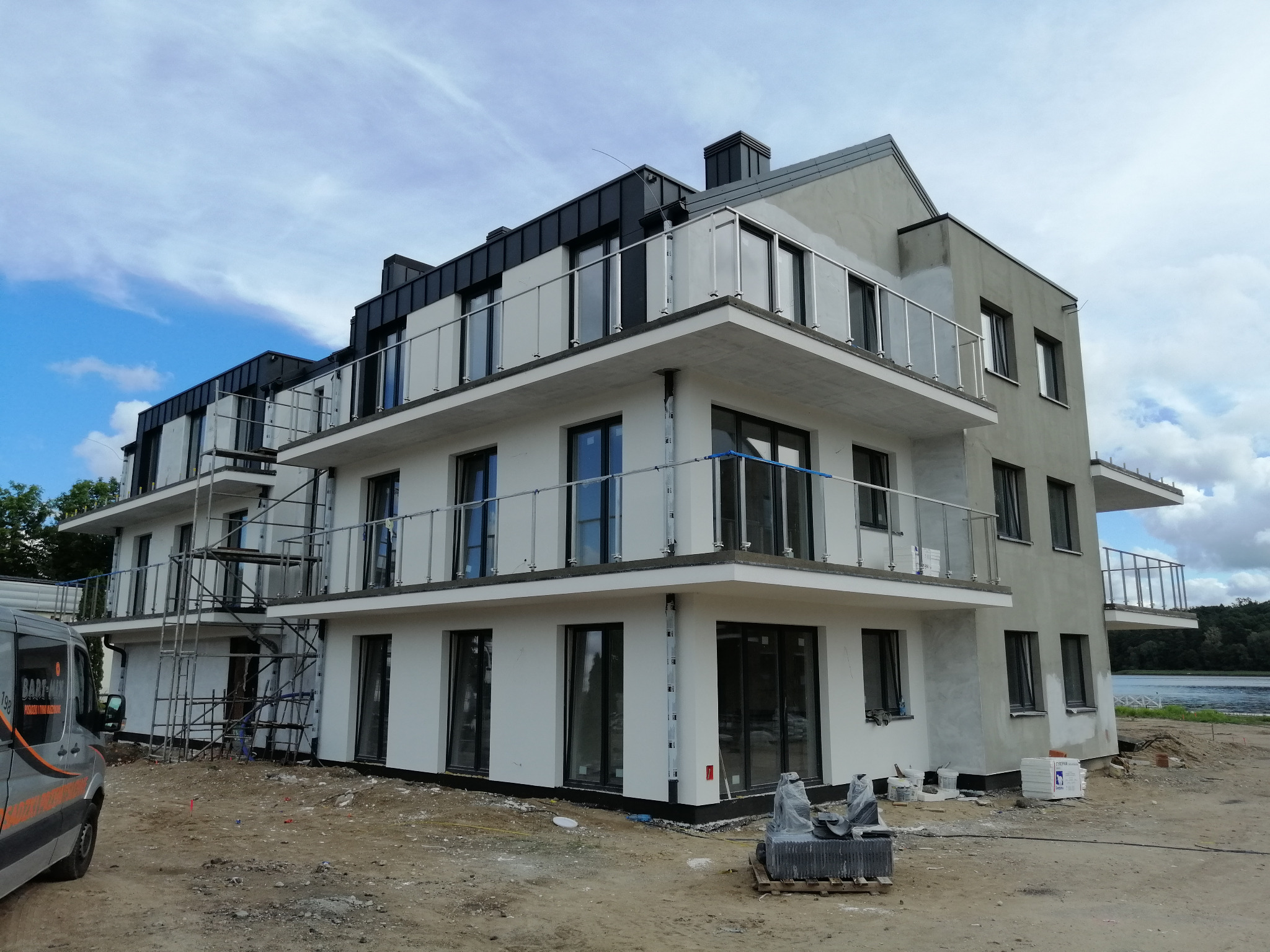 Wielopiętrowy budynek mieszkalny w trakcie wykończenia elewacji, z widocznymi rusztowaniami, balkonami ze szklanymi balustradami i częściowo otynkowaną fasadą w pobliżu jeziora.