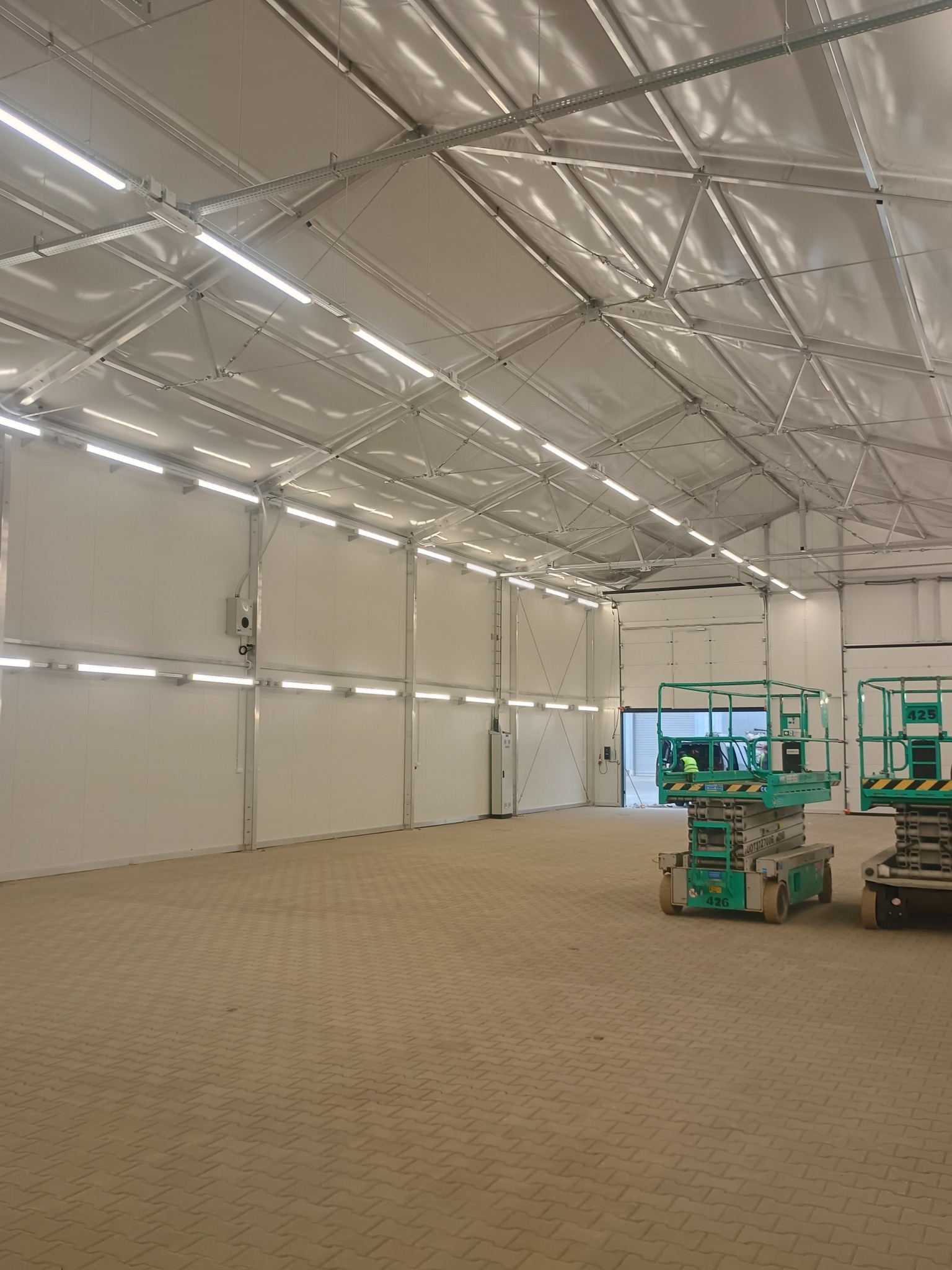 Wnętrze hali z zamontowanym oświetleniem liniowym LED na suficie i ścianach. Dwa podnośniki nożycowe na posadzce z kostki brukowej. Widok na bramę.