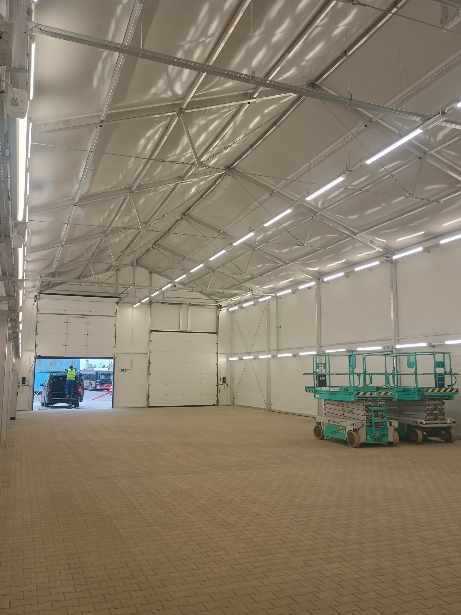 Wnętrze hali z zamontowanym oświetleniem liniowym LED pod sufitem. Widoczny podnośnik koszowy i częściowo otwarta brama wjazdowa z sylwetką osoby.