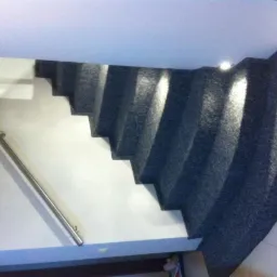 Schody wyłożone szarą wykładziną dywanową, z metalową poręczą po lewej stronie. Widok z dołu na górę, oświetlenie punktowe na ścianie.