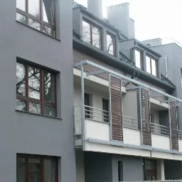 Szara elewacja budynku wielorodzinnego z drewnianymi oknami i ażurowymi przesłonami balkonowymi. W oknach odbija się korona drzewa, a w tle widoczny dach i kominy.