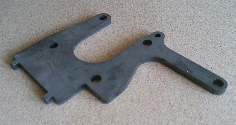 Precyzyjnie wycięty, metalowy element o nieregularnym kształcie z otworami, leżący na beżowym tle. Widoczne delikatne ślady obróbki.