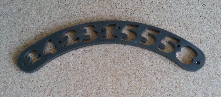 Precyzyjnie wycięty, łukowaty element z ciemnego metalu z ażurowymi cyframi '423:155' leży na korkowej powierzchni. Detal obróbki CNC.