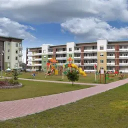 Nowoczesne, wielobarwne bloki mieszkalne z balkonami otoczone zielenią i placem zabaw, sfotografowane w słoneczny dzień.