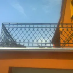 Kuty balustrada balkonowa o dekoracyjnym wzorze rombów, zamontowana na pomarańczowym budynku, widoczna na tle błękitnego nieba.