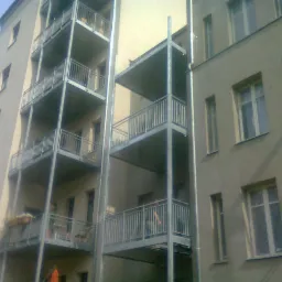 Elewacja budynku z balkonami z metalowymi barierkami. Widoczne pionowe, metalowe elementy konstrukcyjne balkonów.