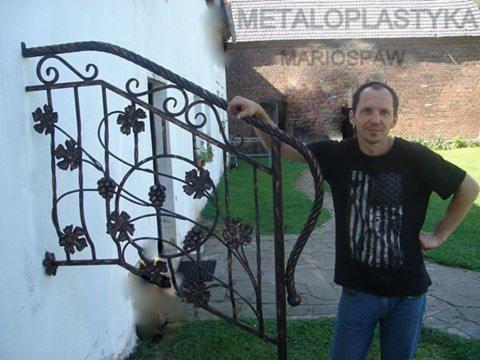 Kute, metalowe zabezpieczenie okienne z motywem winorośli, przymocowane do ściany. Mężczyzna opiera się o konstrukcję. Widoczny trawnik i budynek z cegły w tle.