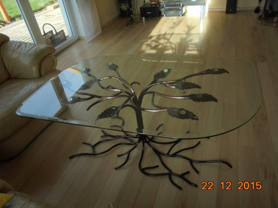 Szklany stolik kawowy z metalową podstawą w kształcie drzewa, stojący na drewnianej podłodze. W tle sofa i okno.