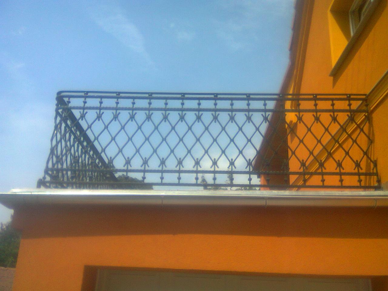 Kuty balustrada balkonowa o dekoracyjnym wzorze rombów, zamontowana na pomarańczowym budynku, widoczna na tle błękitnego nieba.