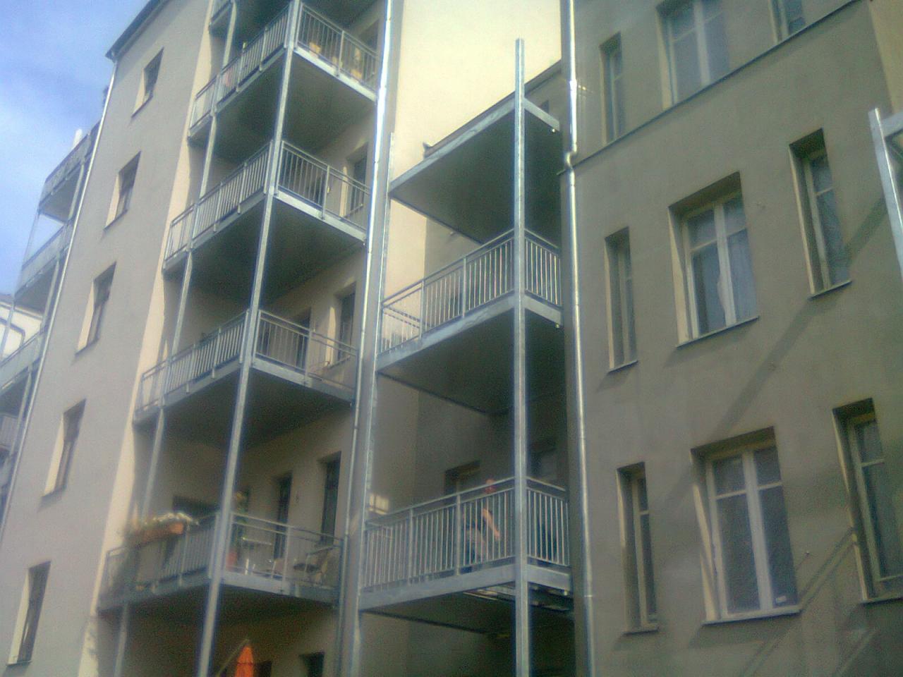 Elewacja budynku z balkonami z metalowymi barierkami. Widoczne pionowe, metalowe elementy konstrukcyjne balkonów.