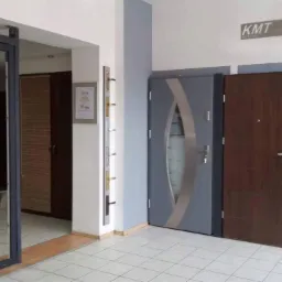 Ekspozycja w salonie z różnymi modelami drzwi zewnętrznych: szklane przesuwne, szare z owalnym przeszkleniem, brązowe z prostokątnym zamkiem i jasne z długim uchwytem.