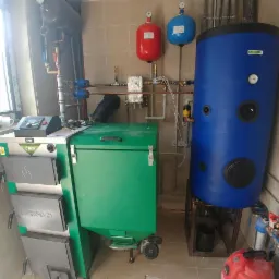 Nowo zainstalowany piec na paliwo stałe Drew-Met z zielonym zasobnikiem i niebieskim zbiornikiem akumulacyjnym, widoczne czerwony i niebieski zbiornik wyrównawczy, całość w pomieszczeniu z beżowymi...