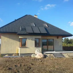 Nowoczesny dom jednorodzinny z panelami słonecznymi na dachu, widok z boku na tle błękitnego nieba, teren wokół domu w trakcie prac ziemnych.