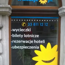 Oklejone okno biura podróży z logo 'wakacje.pl', numerem telefonu i ofertą: wycieczki, bilety lotnicze, rezerwacje hoteli, ubezpieczenia. Żółte słońce.