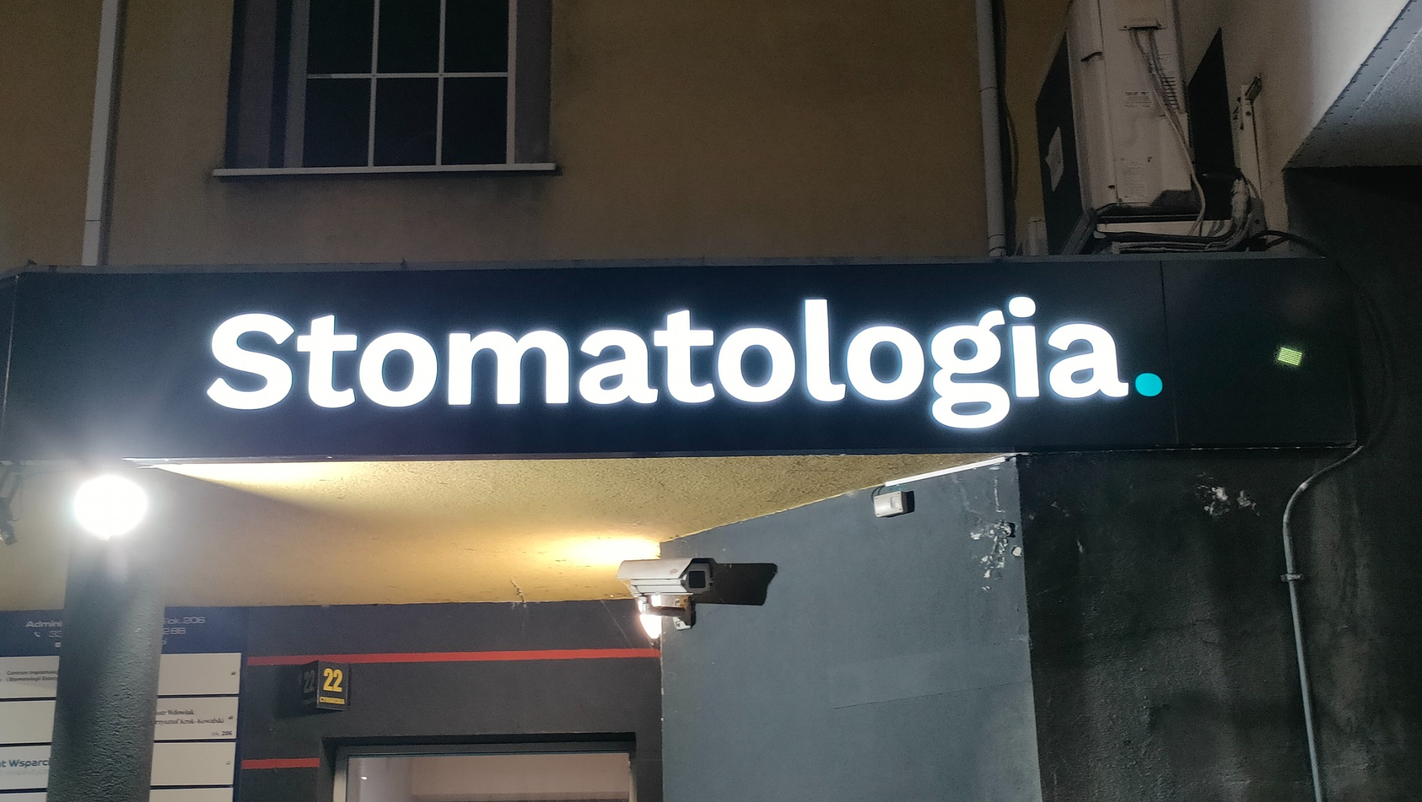 Podświetlany szyld z napisem 'Stomatologia' nad wejściem do budynku. Widoczna kamera monitoringu i numer budynku 22. Szyld w kolorze czarnym z białymi literami.