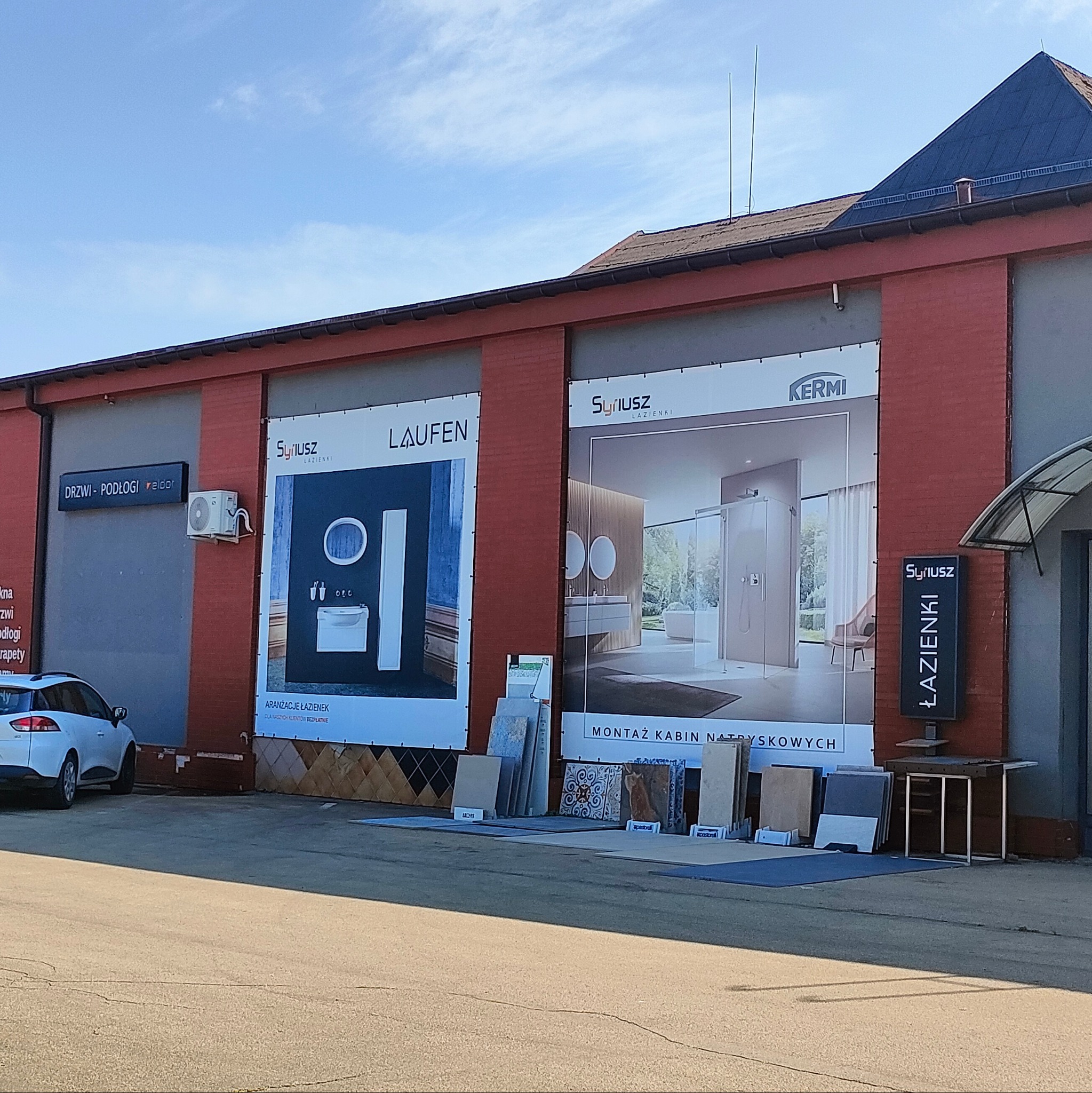 Budynek z banerami reklamowymi łazienek marek Syriusz, Laufen i Kermi. Przed budynkiem ekspozycja płytek ceramicznych. Widoczny fragment zaparkowanego auta.