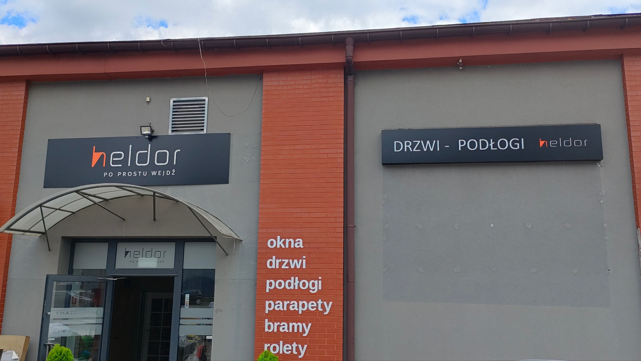 Fasada budynku z szyldami firmy 'heldor' oferującej drzwi i podłogi. Na ścianie wypisane: okna, drzwi, podłogi, parapety, bramy, rolety. Otwarty sklep z wejściem pod zadaszeniem.