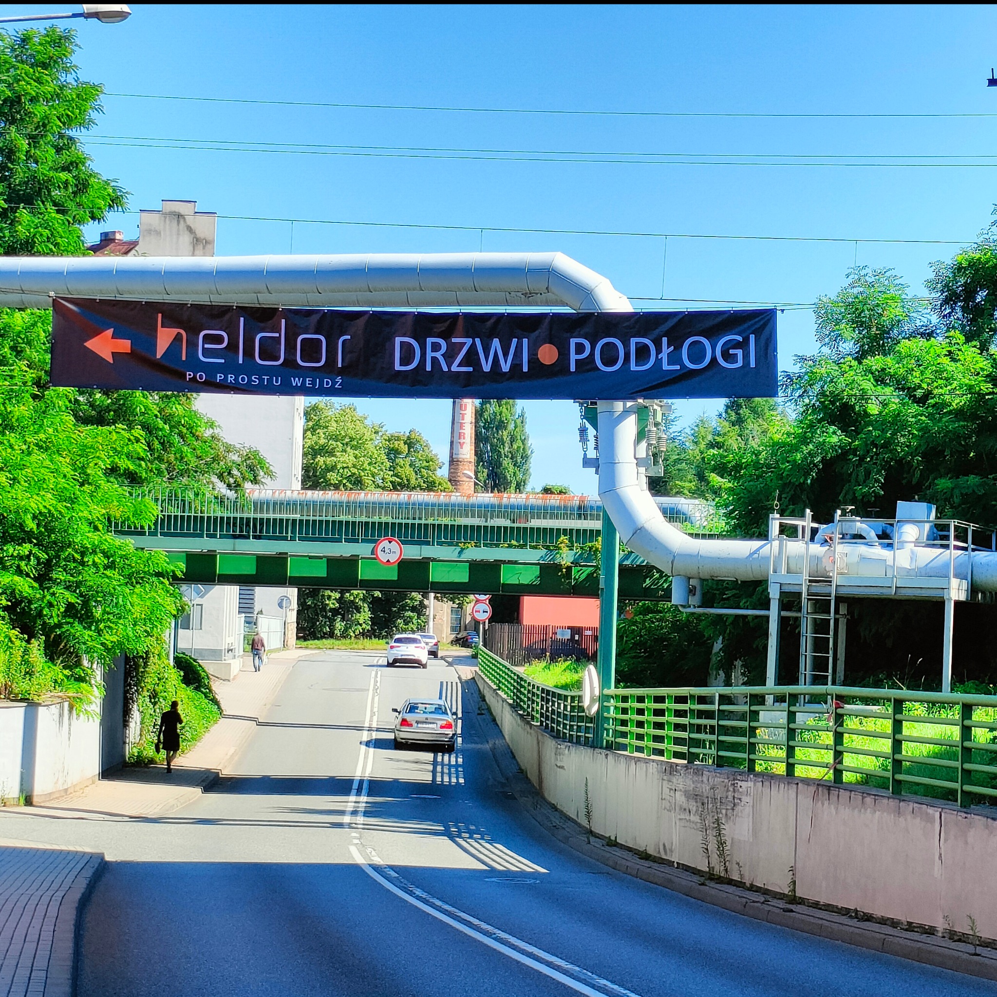 Baner reklamowy firmy Heldor, specjalizującej się w drzwiach i podłogach, zawieszony nad ulicą z przejeżdżającymi samochodami i pieszymi na chodniku.