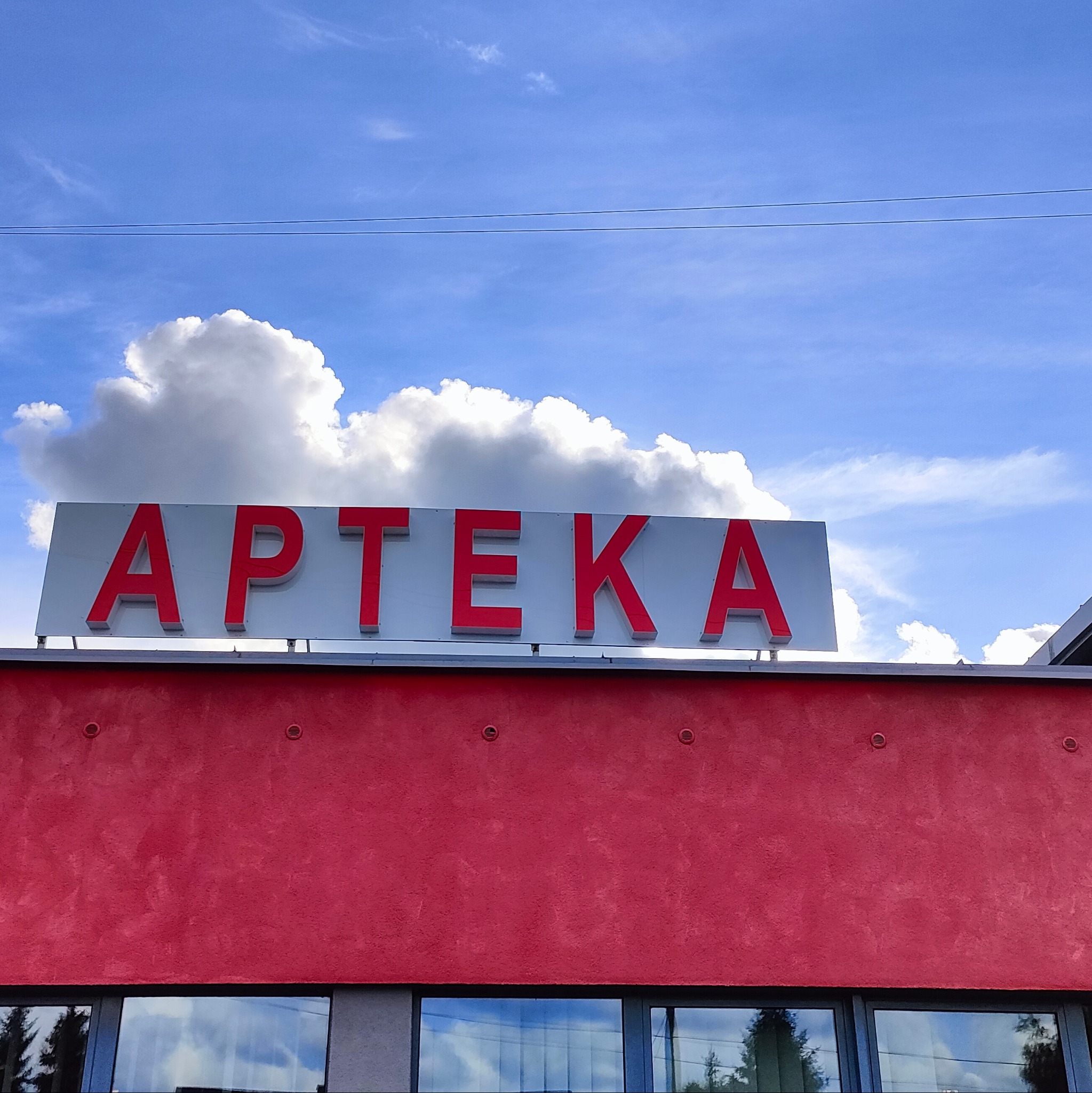 Czerwony budynek apteki z białym szyldem i czerwonym napisem 'APTEKA' na tle błękitnego nieba z chmurami, widziany z dołu.