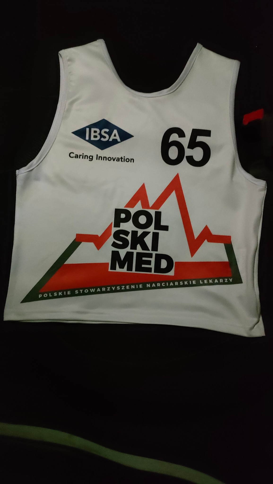 Biała koszulka sportowa z logo IBSA, numerem 65 i napisem POLSKI MED, leżąca na czarnym tle. Koszulka Polskiego Stowarzyszenia Narciarskiego Lekarzy.