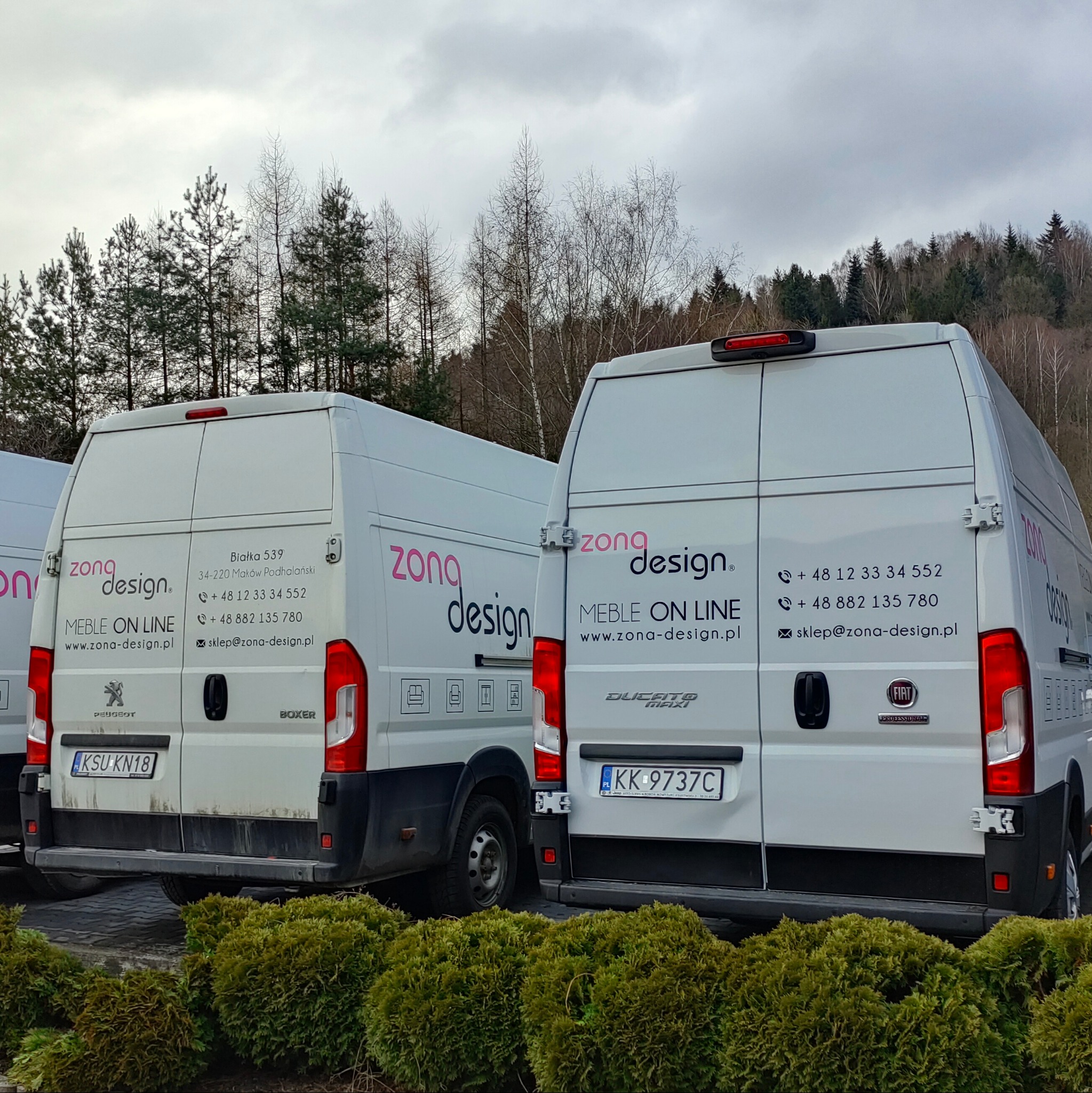Dwa białe busy z różowo-czarnym logo 'zona design' i danymi kontaktowymi, zaparkowane na tle zieleni i drzew. Tablice rejestracyjne widoczne.