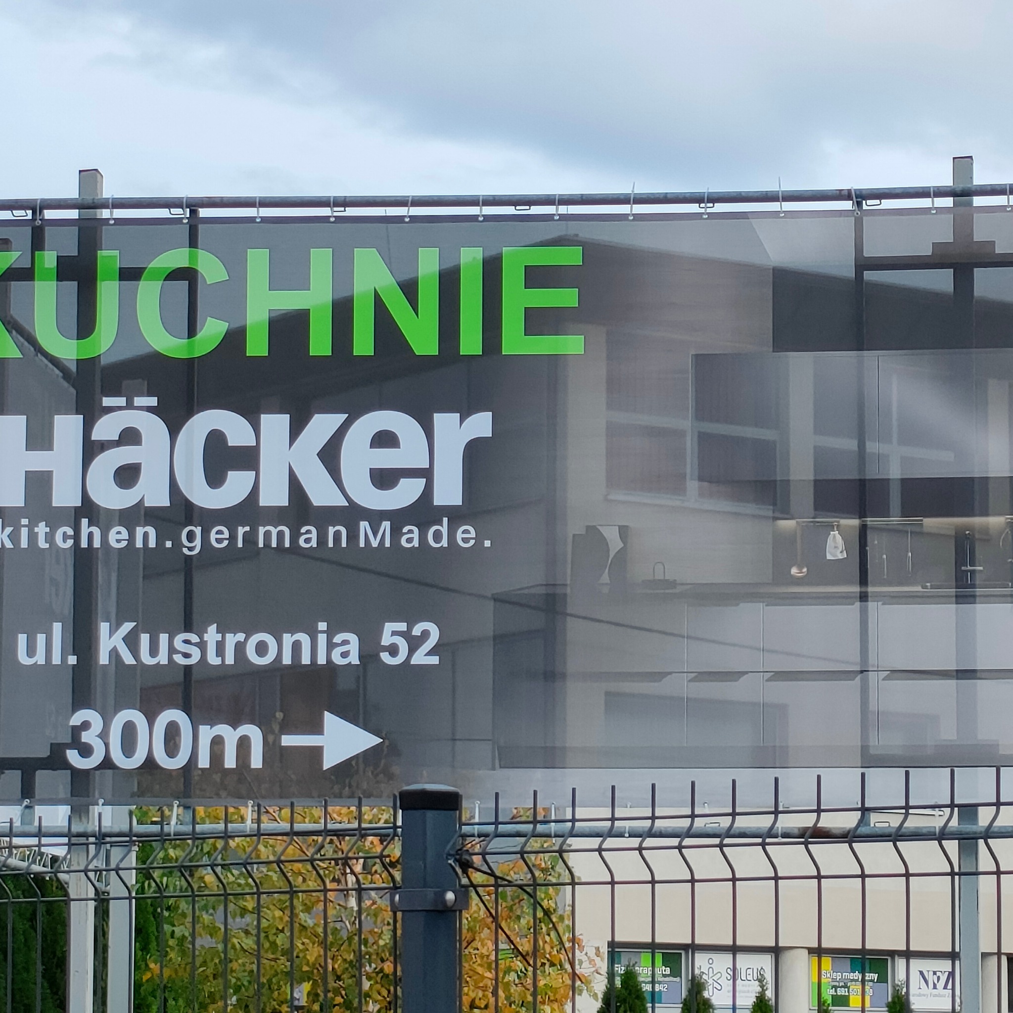 Billboard reklamowy kuchni Häcker, z adresem ul. Kustronia 52 i strzałką wskazującą kierunek, widoczny przez metalowe ogrodzenie, na tle budynku.