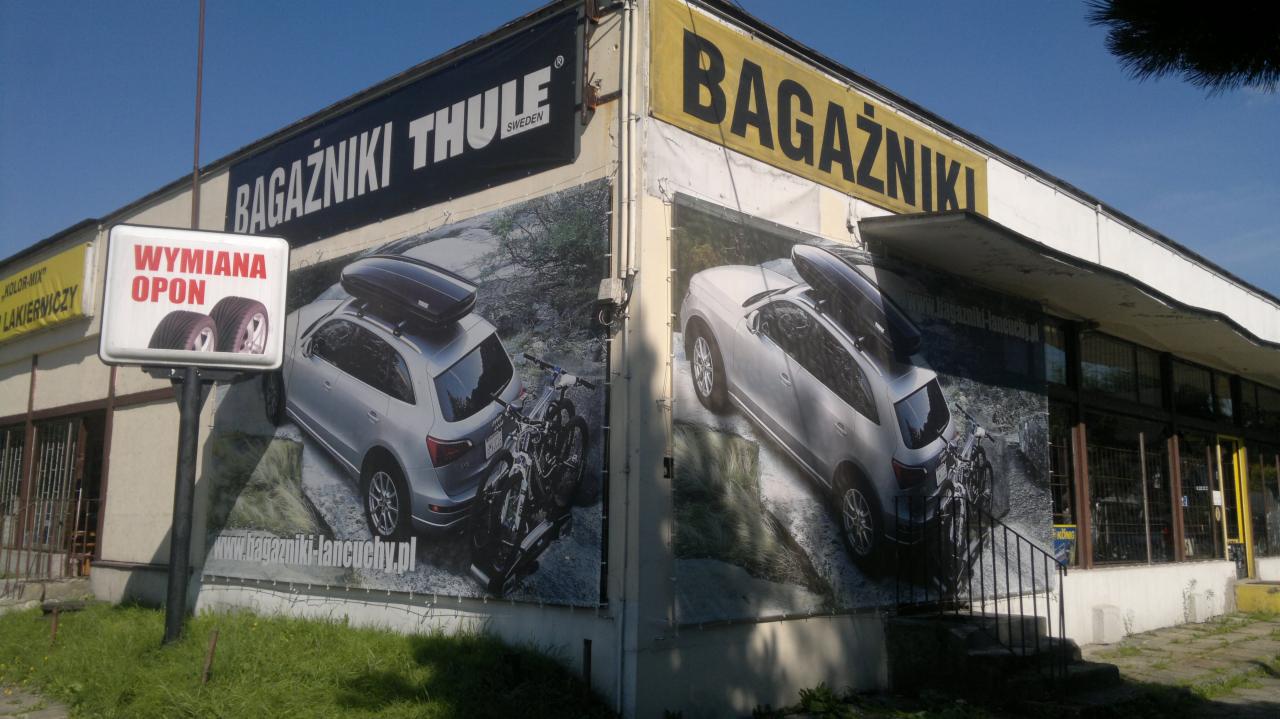 Budynek z reklamą bagażników Thule i billboardem 'Wymiana opon'. Na ścianach auta z bagażnikami dachowymi i motocykl. Widoczny adres strony www.bagazniki-lancuchy.pl.