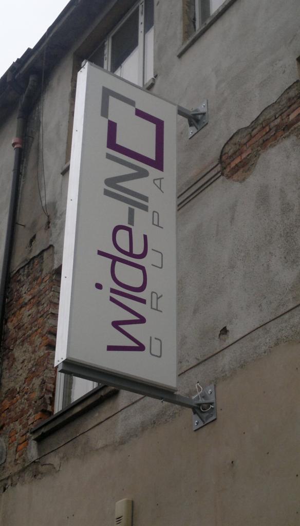 Szyld reklamowy firmy Wide-IN Grupa, zamontowany na elewacji budynku z widocznymi śladami napraw i fragmentami cegieł, w pochmurny dzień.