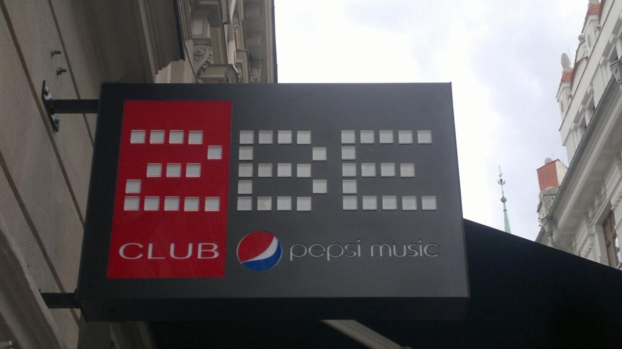 Szyld klubu z napisem 'BE CLUB' i logo Pepsi Music, zamontowany na ścianie budynku. Szary szyld z czerwonym akcentem i białymi kwadratami tworzącymi litery.