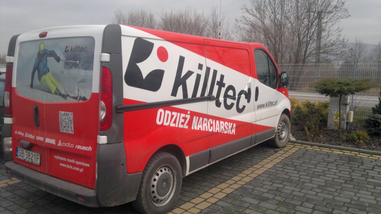 Oklejony folią reklamową samochód dostawczy marki Killtec z grafiką narciarza, zaparkowany na brukowanym parkingu. Czerwono-biała kolorystyka, widoczne logo i hasło 'Odzież Narciarska'.